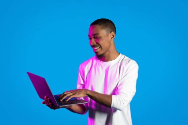 Serbest çalışma, uzaktan çalışma konsepti. Pozitif neşeli, beyaz giyen Afro-Amerikalı adam bilgisayarda klavyeyle yazı yazıyor ve gülümsüyor, online çalışıyor, mavi arka planda, neon ışıkta izole edilmiş, kopyalama alanı