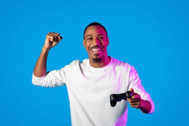 Duygusal olarak mutlu, genç, yakışıklı, beyaz tişörtlü bir adam mavi arka planda video oyunu oynuyor. Neon ışıkta şampiyon, joystick 'i tutuyor ve yumruğunu sıkıyor.
