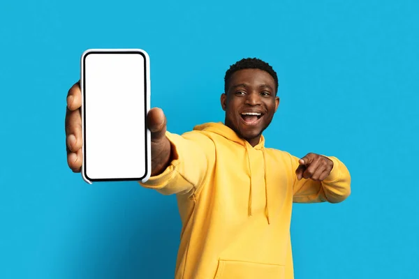 不错的移动应用程序 兴奋而又英俊的黑人年轻人带着模型展示着大大的智能手机 指着白色空白屏幕的小玩意 在蓝色的工作室背景下被隔离开来 — 图库照片