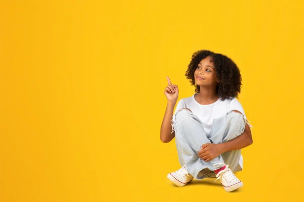身穿白色T恤的快乐的卷曲少年黑人女孩坐在地板上 手指指向空旷的空间 被黄色背景隔离 工作室 教育建议 广告和提供 — 图库照片