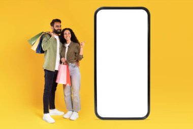 Mobil Alışveriş. Orta Doğulu çift, Smartphone 'a kocaman boş bir ekranla parmak göstererek alışveriş torbaları reklamını sarı stüdyo arkaplanı üzerinden yapıyor. Model