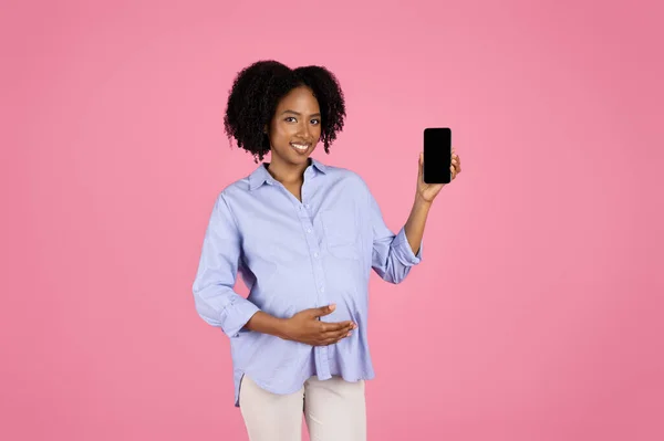 若い妊婦アフリカ系アメリカ人の巻き毛の女性を笑顔でピンクのスタジオの背景に隔離された空白の画面を持つ大きな腹のショースマートフォンでカジュアルに ブログ 期待赤ちゃん アプリ おすすめチャット — ストック写真