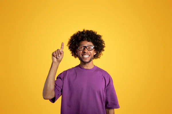 Positive black adult curly man in purple t-shirt and glasses point finger up, isolated on orange background, studio. Got idea, create solution, recommendation, ad and offer