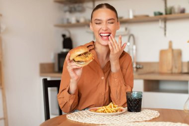 Hamburger yiyen, gülen, zararlı bir yemek yiyen, elinde harika bir burger tutan mutfak masasında oturan heyecanlı genç bayan.