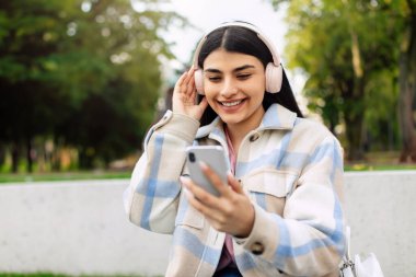 Mutlu İspanyol bayan akıllı telefon kullanıyor ve kulaklıkla müzik dinliyor, internette mesaj atıyor ve sörf yapıyor, parkta oturuyor, bedava fotokopi çekiyor.