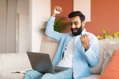 Öyle mi? Modern oturma odasında oturan neşeli Orta Doğulu Adam dizüstü bilgisayar kullanıyor, yumruklarını sallıyor ve sevinç içinde yumruklarını sallıyor. Online Haberler ve Şans 'ı kutlayan Hintli adam evde 