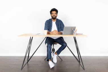 Mutlu, yakışıklı, sakallı, genç Hintli bir adam ev ofisinin çalışma masasında oturuyor, beyaz duvar altyapısı üzerinde, dizüstü bilgisayar kullanıyor, kahve içiyor, doğu milenyumu adamı online çalışıyor, uzayı kopyalıyor.