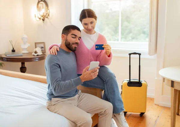 銀行の申し出だ 観光客のカップル予約クレジットカードと携帯電話を使用してホテルの部屋はベッドルームの屋内に座っています 旅行者アプリケーションでオンラインショッピング 簡単かつ安全な支払いオファー — ストック写真