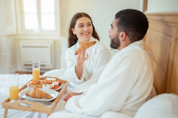 朝が好き ベッドでの朝食中に夫にクロワッサンを提供する楽しい妻は ホテルのベッドルームのインテリアで食べ物のトレイでお互いの会社を楽しんでいます 新婚旅行の休暇の喜び — ストック写真