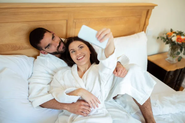 新婚旅行の瞬間 幸せな多民族のカップルは 電話を使用して自撮りのためにポーズをとって 抱っこし ホテルのスイートのインテリアでベッドに横たわって幸せを捉えます 新婚旅行中にガジェット楽しい — ストック写真