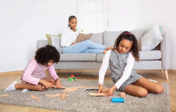 stock image Children Pastime. Black Preteen Daughters Playing Together With Toy Railway While Mother Reading Book Resting On Sofa In Living Room Interior. Selective Focus On Kids Girls