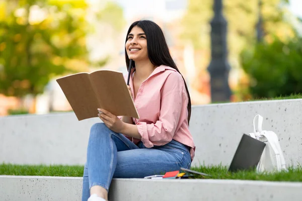 ノートパソコンで公園に座っている間にノートにペン書きを保持しているかなり幸せなヒスパニック系の女性 夢のような女性の思考 将来を計画 — ストック写真