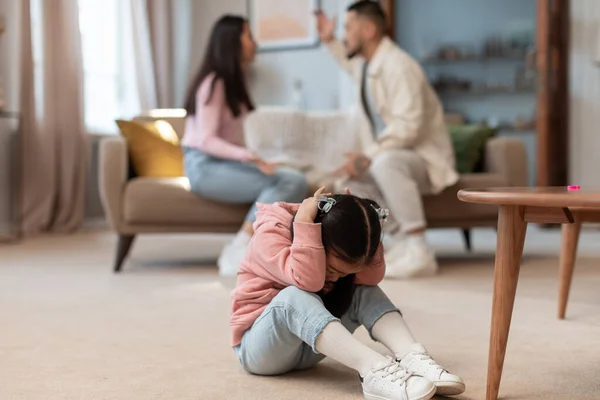 Family quarrels and relationship problems. Asian baby girl covering ears while her angry parents fighting on the background, depressed child feeling lonely in living room at home. Selective focus