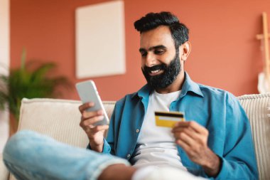 Çevrimiçi Mobil Alışveriş. Kredi kartı ve cep telefonu olan mutlu Hintli adam koltukta oturuyor, internet siparişini onaylıyor, ev içi uygulamasını kullanıyor. Guy Smartphone aracılığıyla Web mağazasından bir şey satın alıyor