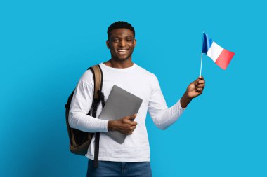 Sırt çantası taşıyan neşeli, yakışıklı, genç, siyah bir adam. Öğrenci bilgisayarı, Fransa bayrağı, mavi arka planda izole edilmiş. Yurtdışında eğitim, Avrupa konsepti eğitimi, kopyalama alanı