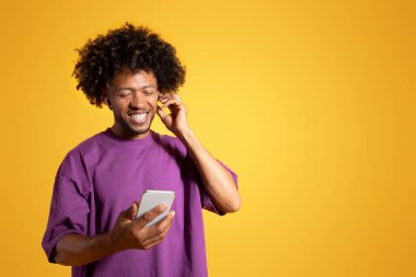 Mor tişörtlü, kablosuz kulaklıklı neşeli siyah kıvırcık adam telefona bakıyor, turuncu arka planda, stüdyoda izole edilmiş. Rahatlamak için müzik dinle, ses uygulaması, reklam ve teklif