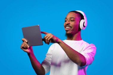 Neşeli, mutlu, yakışıklı, beyaz gömlek giyen, kablosuz kulaklık ve dijital ped kullanan, tablet ekrana dokunan, güzel içeriği internetten izleyen, mavi arka plan, kopyalama alanı olan siyah bir adam.