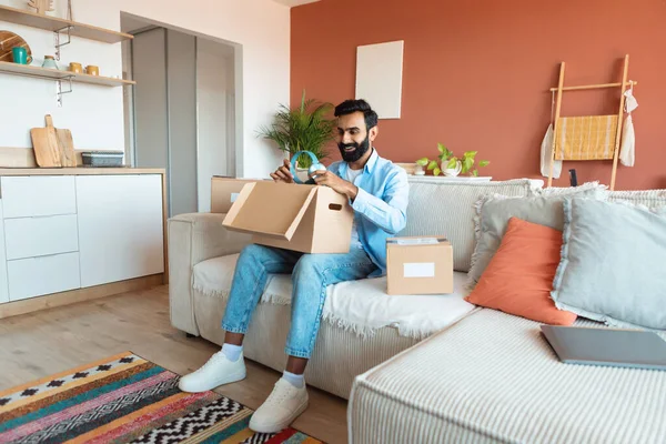 買い物と配達 購入されたヘッドフォンを受信し 屋内でオンライン注文を得る 陽気なインドの男はカートン小包を開きます 顧客の男アンパック段ボール箱に座ってソファに家に — ストック写真