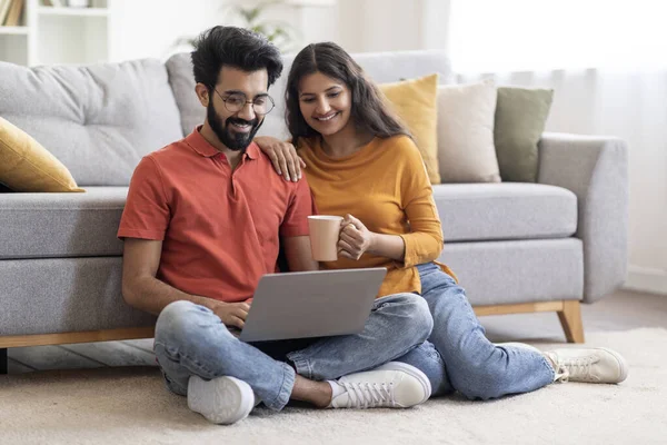 自宅のリビングルームでノートパソコンとコーヒーリラックスした笑顔のインドのカップル 幸せな若い東の配偶者が床に休んで 一緒にコンピュータを使用して オンラインショッピングやインターネットの閲覧 — ストック写真