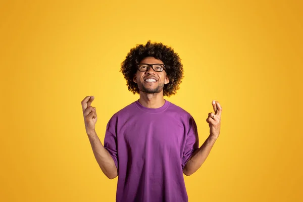 快乐的黑色卷曲男子 身穿紫色T恤和眼镜 做交叉手指手势 隔离在橙色背景下 工作室 希望赢 希望广告和报价 — 图库照片