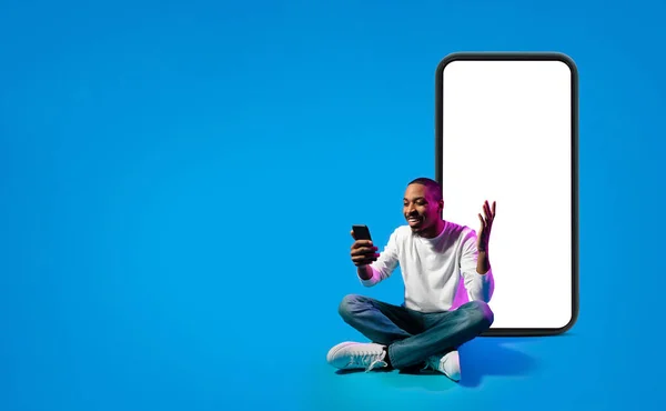 兴奋而又英俊的年轻黑人男子穿着休闲装 用智能手机和手势 坐在地板上的巨大的电话 蓝色背景的白色空白屏幕在霓虹灯下 复制空间 — 图库照片