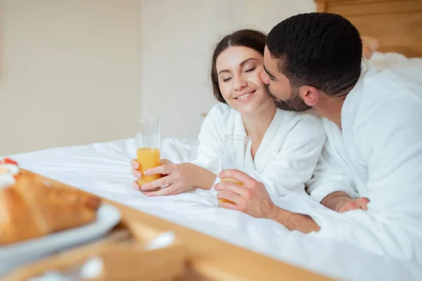 甜蜜的蜜月 丈夫亲吻妻子就像爱夫妻享受橙汁 躺在旅馆房间里的床上 配偶在室内早餐时放松分享浪漫假期的时刻 — 图库照片