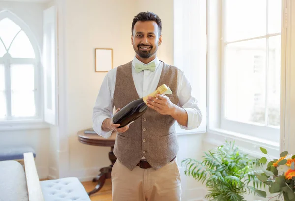 Smiling Indian Waiter Guy With Bottle Of Sparkling Wine Ready To Serve Alcohol Drink, Standing At Luxury Hotel Room Interior, Wearing Uniform. Professional Sommelier Career