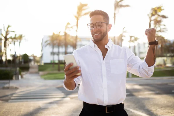 喜びに満ちたヨーロッパの若いビジネスマン携帯電話を保持し 拳を振る 男起業家ジェスチャーはい成功を祝う 偉大なニュースを読む 屋外に立つ — ストック写真