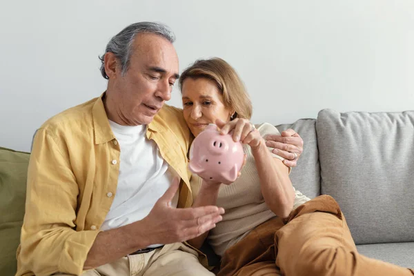 財政難だ 高齢者のカップルは お金のない空の貯金箱をチェックし 経済危機に苦しんで 自宅のソファの上に フリースペースに座って動揺 財政難に直面している退職者のカップル — ストック写真