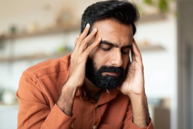 Baş ağrısı ve stres. Evde koltukta otururken şakaklara dokunan Hintli adam baş ağrısı, depresyon ya da yakın çekim portresi yüzünden acı çeken genç sakallı adam.