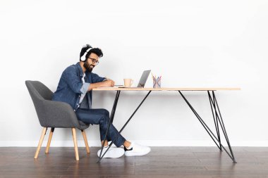 Neşeli, yakışıklı, genç, ortadoğulu bir adam, dizüstü bilgisayar, kablosuz kulaklık kullanarak, not alarak masa başında oturuyor, milenyum adamı online eğitime katılıyor, evden giyiniyor, beyaz duvar arka planı, kopyalama alanı.