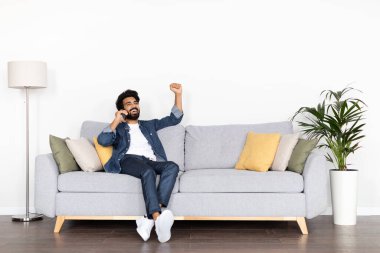 Heyecanlı, genç, yakışıklı, orta doğulu, günlük kıyafetler giyen, oturma odasında koltukta oturan, evde telefon görüşmesi yapan, el kol hareketi yapan ve ünlenen, iyi haber: