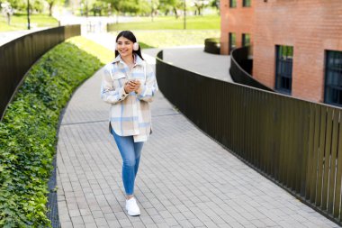 Mutlu Latin bayan öğrenci cep telefonu kullanıyor ve gülümsüyor, dışarıda yürüyor ve kablosuz kulaklıkla müziğin keyfini çıkarıyor, kampüste dersten sonra mola veriyor, boş yer var.