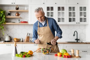 Gülümseyen kıdemli adam mutfakta taze sebze salatası pişiriyor, zeytinyağı ve baharat katıyor, önlük giymiş yaşlı beyefendi evde sağlıklı vejetaryen yemekleri yapıyor, uzayı kopyalıyor.