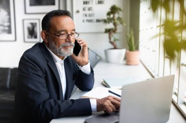 Avrupalı kıdemli takım elbiseli güler yüzlü bir işadamı ofis içi masasındaki akıllı telefondan dizüstü bilgisayarlı aramalar yapan gözlüklü. Uzaktan çalışma, iş uygulaması, iletişim ve teknoloji toplantısı