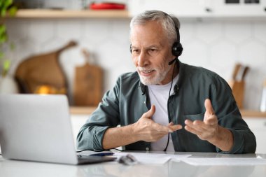 Mesafe iletişimi. Kulaklık takmış yakışıklı bir adam mutfak masasında otururken video çekiyor, yaşlı bir erkek internet üzerinden iletişim kuruyor, bilgisayar ve kulaklık kullanıyor.