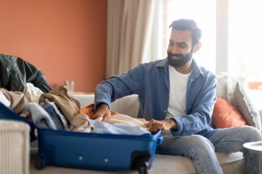 Tatil kıyafetleri. Gülümseyen Hintli adam seyahat çantalarını topluyor, erkek kıyafetlerini evdeki kanepeye oturtuyor. Sakallı Orta Doğulu Adam, Bavulları Kapalı Tur Gezisine Hazırlıyor