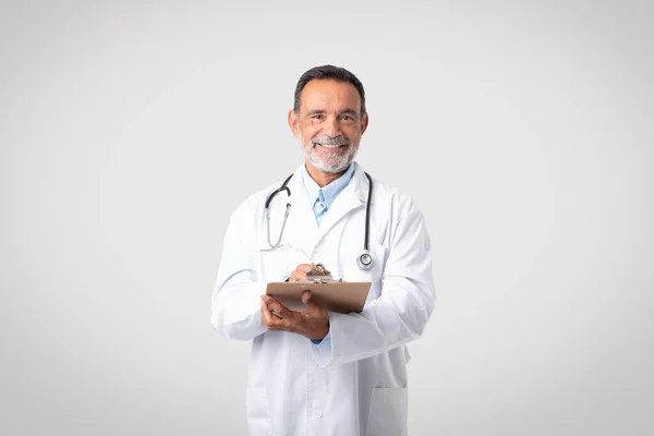 白いコートを着た幸せな白人の成熟した男性医師は グレーの背景 スタジオに隔離されたタブレットでノートを作ります 生命保険 ヘルスケア 広告と提供 クリニックでの医療 — ストック写真