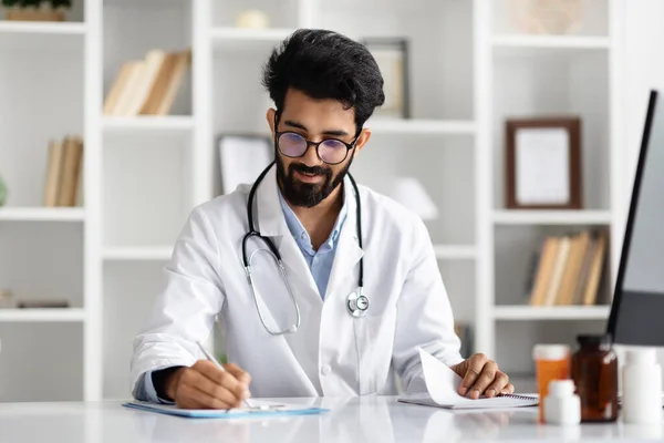 中东英俊的年轻人 身穿医疗外套 脖子上戴着听诊器的眼镜 在现代诊所的办公室里工作 开处方 复印空间 — 图库照片
