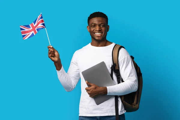 穿着休闲装背着背包 拿着笔记本电脑 拿着英国国旗 身穿蓝色背景衣服的英俊黑人男子笑着 留学教育 留学观念 复制空间 — 图库照片
