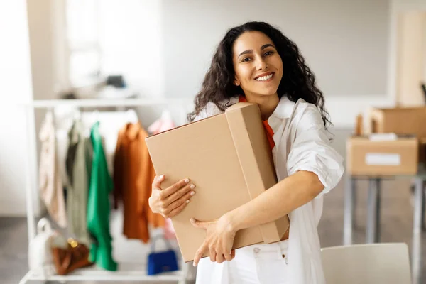 stock image Clothing Store Owner Woman Hugging Cardboard Box With Trendy Clothes Posing In Fashionable Showroom, Smiling To Camera. Happy Entrepreneur Embracing Her Fashion Business Success