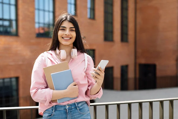 スマートフォンを持つラテン語の陽気な学生の女性の肖像画や 屋外でポーズをとってカメラで笑顔 大学の近くに立っている間に電話を使用して女性 — ストック写真