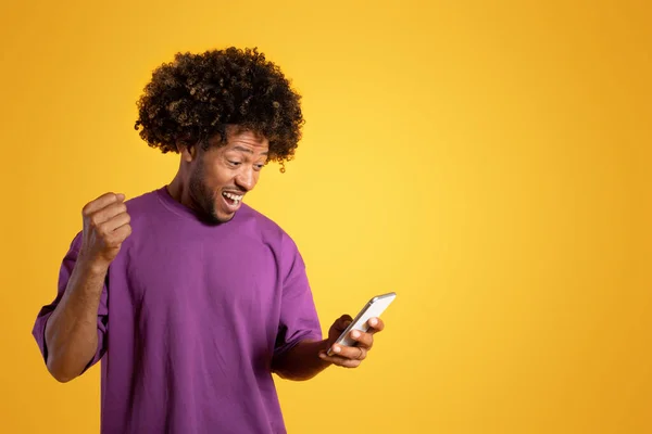 보라색 티셔츠를 곱슬머리의 남자가 스마트폰을 성공을 축하하며 오렌지 스튜디오에 고립되어 — 스톡 사진