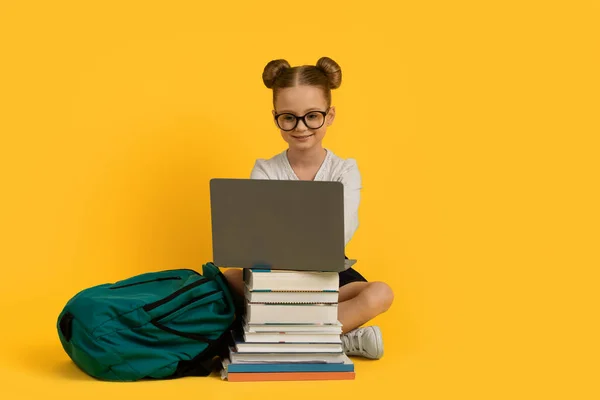 オンライン学習コンセプト 黄色いスタジオの背景の上のブックスタックに座っている間 ノートパソコンで宿題をしているかわいい女の子 笑顔のPreteen女性の子供は リモートレッスンを楽しんで スペースをコピーします — ストック写真