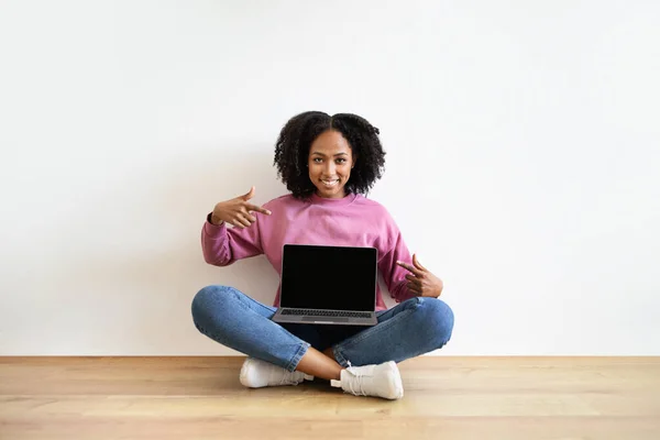 正の千年黒の女性が床に座って 白い壁の背景に部屋のインテリアに空白の画面をラップトップで指をポイントします ウェブサイトの勧告とアドバイス オンラインサービス 広告やオファー — ストック写真