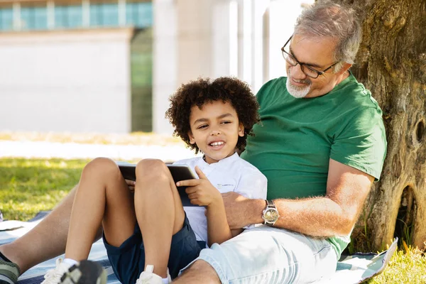 stock image Cheerful senior man with beard and black little boy hugging, have fun, relax in park, watch video on tablet, outdoor. Relationship, grandson and grandfather, free time and walk, chat, game