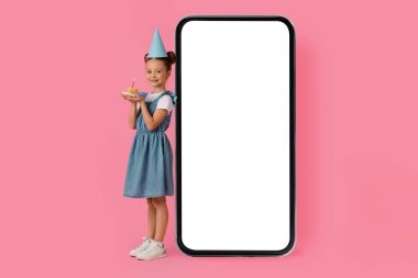 Doğum günü pastası olan şirin küçük kız Büyük Boş Akıllı Telefon 'un yanında, Beyaz Ekranlı Neşeli Genç Kız Çocuk Doğum gününü kutluyor ve Yeni Mobil Uygulama Öneriyor, Mobil Mobil Uygulama,