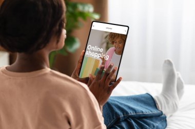 Afro-Amerikalı kadın evde moda mağazası olan tablet kullanıyor, yeni kıyafetler arıyor, cihazla online alışverişin tadını çıkarıyor. Moda mağazası için web sitesi ya da uygulama