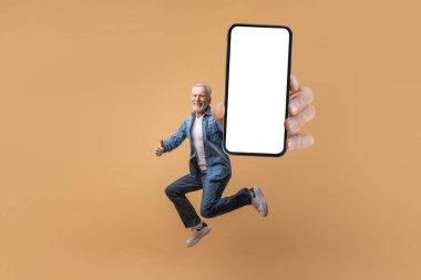 E-ticaret, perakende, harika bir teklif. Heyecanlı, gri saçlı, son sınıf öğrencisi elinde beyaz ekranı, bej arka planı, baş parmağını gösteren, taklit eden, uzayı kopyalayan kocaman bir akıllı telefonla havaya atlıyor.