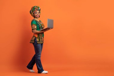 Çevrimiçi asistan, sohbet robotu. Mutlu, çekici, Afrikalı kostümlü, elinde bilgisayar bilgisayarıyla dolaşan, turuncu stüdyo arka planında izole edilmiş siyah bir kadın.